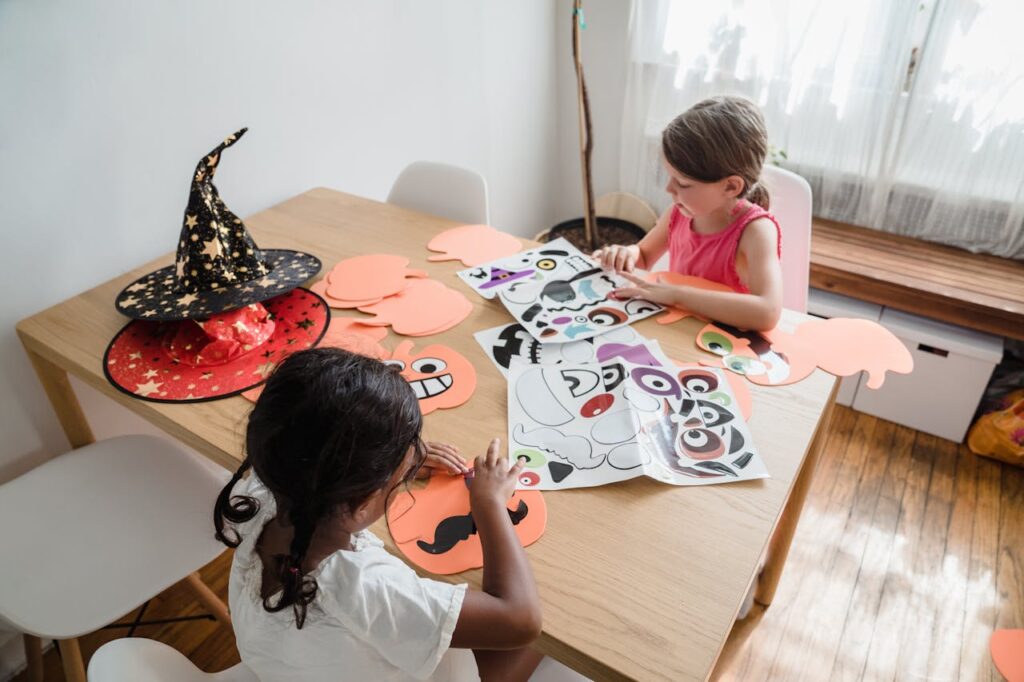 Two kids making Halloween crafts at a table with paper and witch hat.